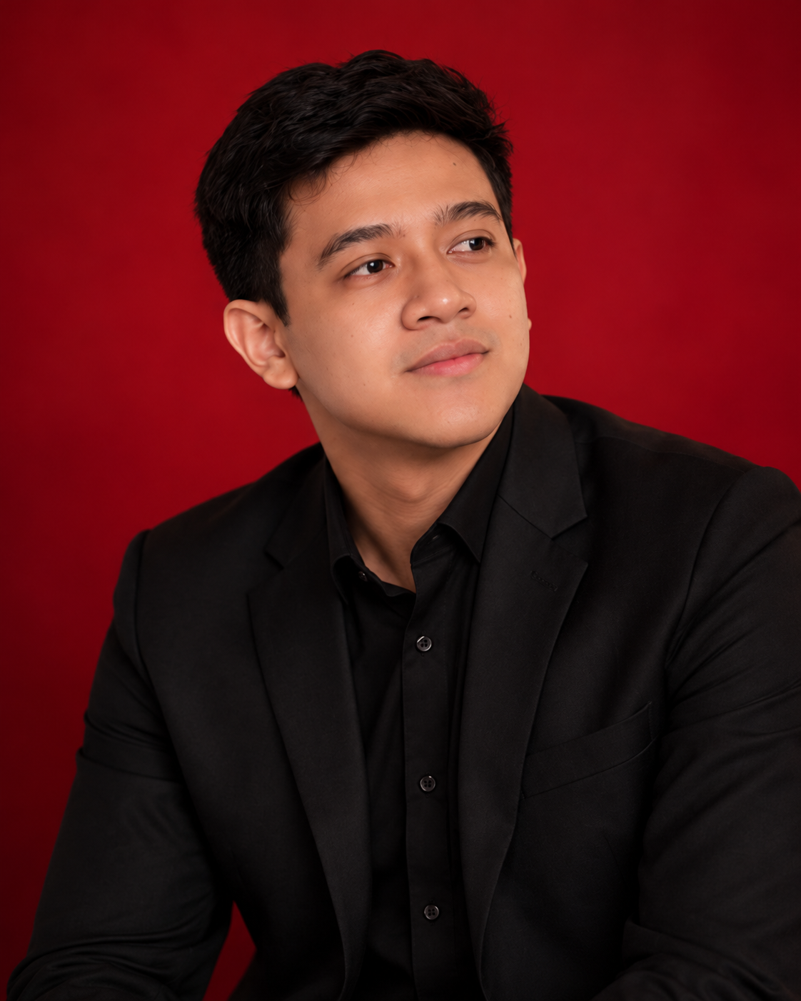 Andrew Bajar in a black suit, looking to the side against a burgundy studio background.
